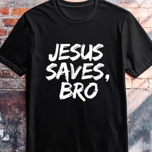 Op de afbeelding: Zwart T-shirt met de witte tekst "JESUS SAVES, BRO" in een gedurfde, handgeschreven stijl. Het shirt heeft een ronde hals en lijkt gemaakt van een zacht materiaal. De merknaam "DISCIPLE Ink" staat rechtsboven gedrukt.