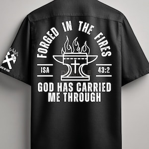 Puede incluir: Camisa negra con texto y gráficos blancos. La parte posterior presenta las palabras "FORGED IN THE FIRES" alrededor de un diseño de yunque y cruz, con "ISA 43:2" y "GOD HAS CARRIED ME THROUGH". La manga izquierda tiene un gráfico blanco.