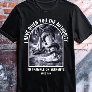 May include: Black t-shirt with a graphic of a foot stepping on a snake, with the text "I HAVE GIVEN YOU THE AUTHORITY TO TRAMPLE ON SERPENTS" and "LUKE 10:19". The shirt also has the text "DISCIPLE ink".