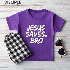 Op de afbeelding: Een paars T-shirt met de tekst "JESUS SAVES, BRO" in het wit. Daarnaast een zwart-wit geruite broek en een hartvormige zonnebril.