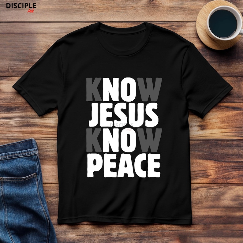 May include: Black t-shirt with the text "KNOW JESUS KNOW PEACE" in white and gray lettering. The shirt is displayed on a wooden surface with a cup of coffee and a pair of blue jeans.