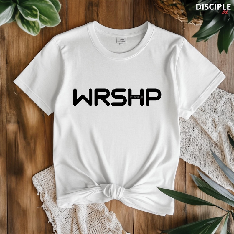 May include: A white t-shirt with black text that reads 'WRSHP' with the word 'DISCIPLE' in smaller text below. The t-shirt is knotted in the front and is laying on a wooden surface with green plants in the background.