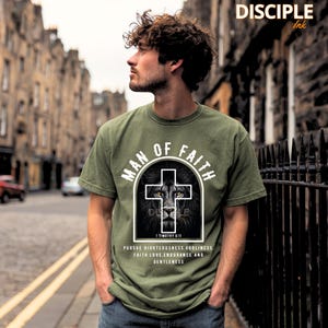 May include: Olive green t-shirt with the text "MAN OF FAITH" in a circular design. A lion's face is centered within a white cross. The text "1 TIMOTHY 6:11" and other words are below the cross. The brand name "DISCIPLE Ink" is at the top.