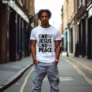 May include: White t-shirt with the text "KNOW JESUS KNOW PEACE" in gray and black. The shirt is short-sleeved and worn by a person in light blue jeans. The image is taken outdoors on a city street.
