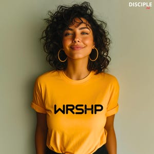 May include: A woman wearing a yellow t-shirt with the text "WRSHP" in black. The text is in a bold, sans-serif font. The woman has curly brown hair and is wearing gold hoop earrings.