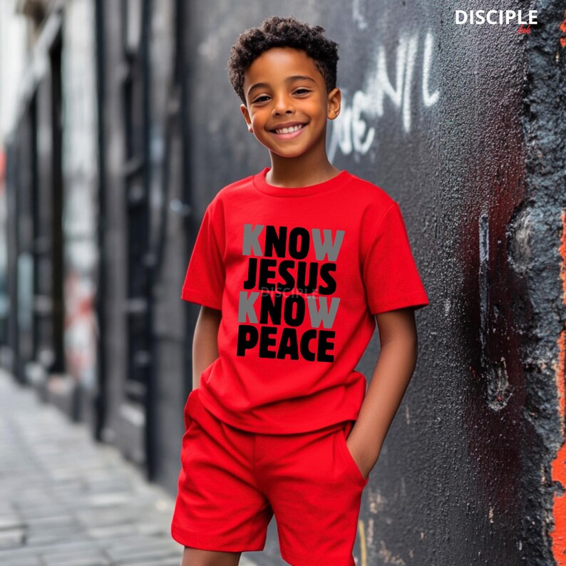 May include: A red t-shirt and shorts set. The t-shirt features the text "KNOW JESUS KNOW PEACE" in a mix of gray and black lettering. The word "DISCIPLE" is printed in white in the upper right corner.