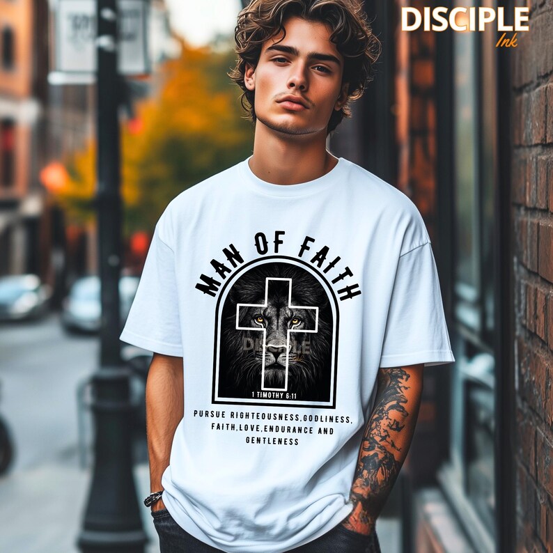 May include: White t-shirt with a graphic design featuring a lion's face within a cross, the words "MAN OF FAITH" and text from 1 Timothy 6:11. The shirt also includes the words "PURSUE RIGHTEOUSNESS, GODLINESS, FAITH, LOVE, ENDURANCE AND GENTLENESS".