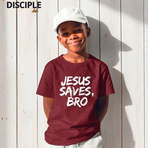 Op de afbeelding: Een bordeauxrood t-shirt met de tekst "JESUS SAVES, BRO" in het wit, gedragen door een persoon met een witte pet. Het shirt heeft een ronde hals en korte mouwen. De achtergrond is een lichte houten muur.