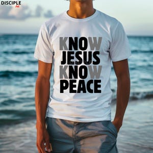 May include: White t-shirt with the text "KNOW JESUS KNOW PEACE" in gray and black block letters. The shirt is a crew neck style and is being worn in front of a blurred ocean background.