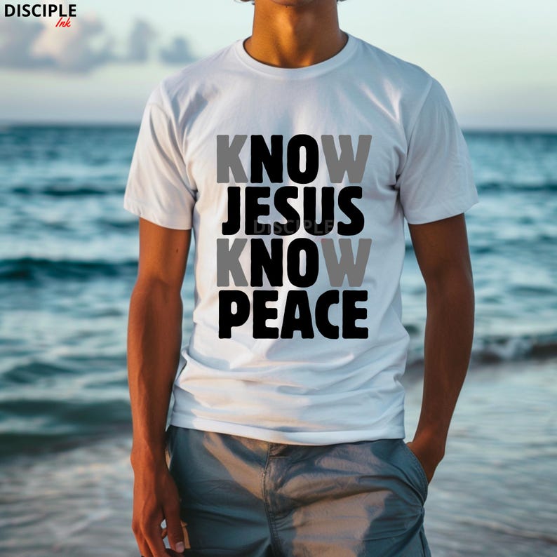 May include: White t-shirt with the text "KNOW JESUS KNOW PEACE" in gray and black block letters. The shirt is a crew neck style and is being worn in front of a blurred ocean background.