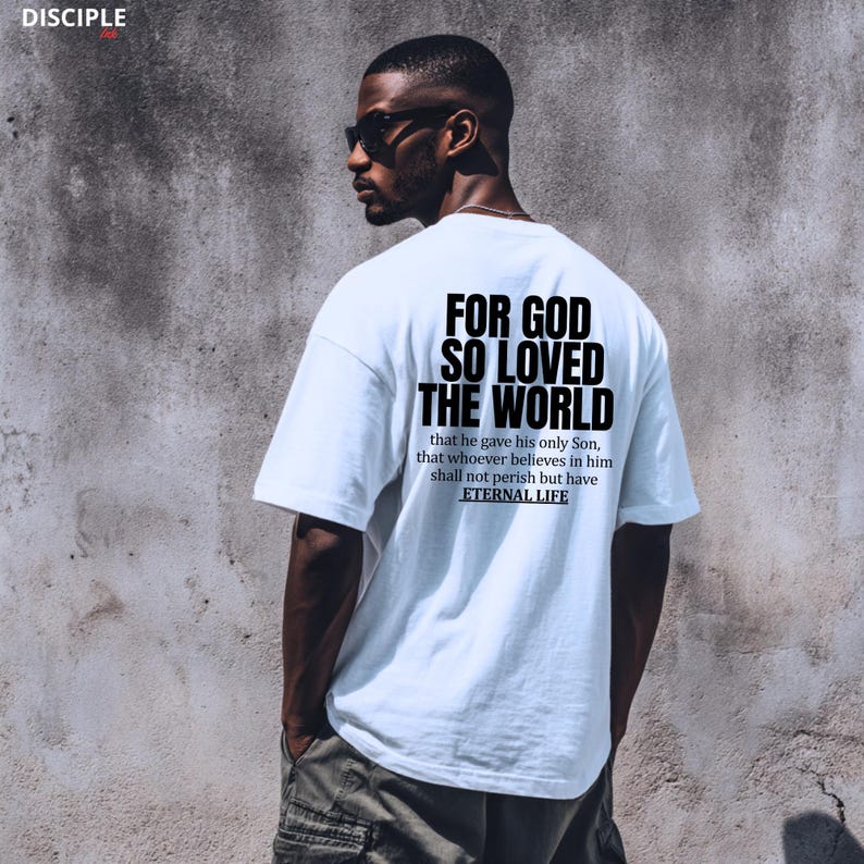 Pode incluir: Camiseta branca com o texto "FOR GOD SO LOVED THE WORLD" e um vers&iacute;culo b&iacute;blico nas costas. A camiseta tem gola redonda. O texto est&aacute; em negrito, com uma fonte sem serifa.