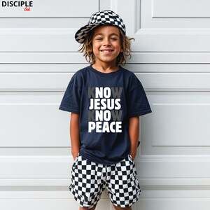 May include: A child wearing a navy blue t-shirt with the words "KNOW JESUS KNOW PEACE" in white and gray. The child is also wearing black and white checkered shorts and a matching baseball cap. The child is smiling.