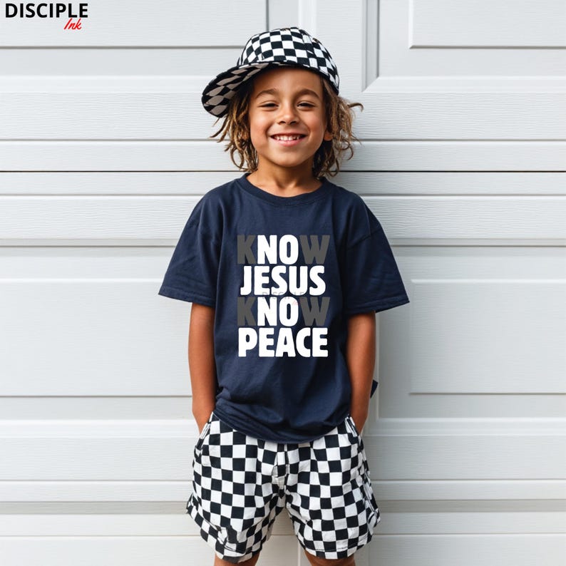 May include: A child wearing a navy blue t-shirt with the words "KNOW JESUS KNOW PEACE" in white and gray. The child is also wearing black and white checkered shorts and a matching baseball cap. The child is smiling.