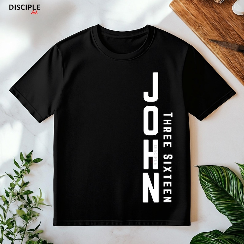 Pode incluir: Camiseta preta com o texto branco "JOHN THREE SIXTEEN" alinhado verticalmente &agrave; direita. A marca "DISCIPLE Ink" est&aacute; no canto superior esquerdo. A camisa &eacute; exibida em uma superf&iacute;cie de madeira com folhagem verde.