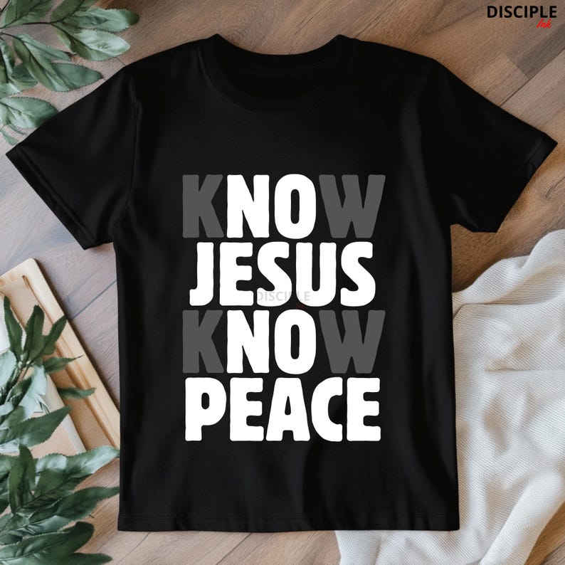 May include: Black t-shirt with the text "KNOW JESUS KNOW PEACE" in white and gray block letters. The shirt is a crew neck style and appears to be made of a soft material. The shirt is displayed on a light-colored surface.