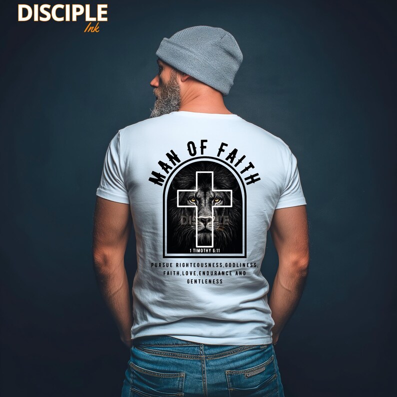 May include: White t-shirt with the text "MAN OF FAITH" above a cross design featuring a lion's face. The text "1 TIMOTHY 6:11" and other words are printed below the cross. The shirt is worn by a person wearing a gray beanie and blue jeans.