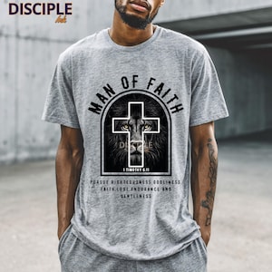 May include: A gray t-shirt with the text "MAN OF FAITH" above a cross design with a lion's face. Below the design is the text "1 TIMOTHY 6:11" and a quote about faith. The shirt is a crew neck.