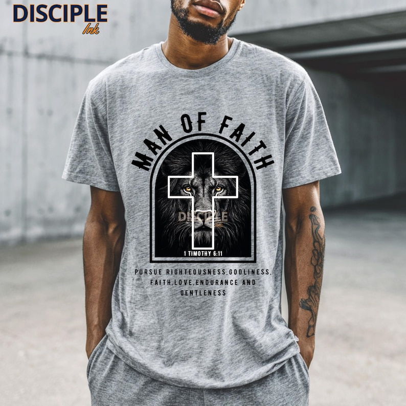 May include: A gray t-shirt with the text "MAN OF FAITH" above a cross design with a lion's face. Below the design is the text "1 TIMOTHY 6:11" and a quote about faith. The shirt is a crew neck.