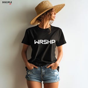 May include: A woman wearing a black t-shirt with the word "WRSHP" printed in white on the front. She is also wearing denim shorts and a straw hat.