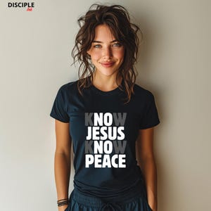 May include: A navy blue t-shirt with the words "KNOW JESUS KNOW PEACE" in a stacked design. The top text is white, the middle text is gray, and the bottom text is white. The shirt is short-sleeved.