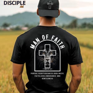 May include: Black t-shirt with a white graphic of a lion inside a cross, the words "MAN OF FAITH" and "1 Timothy 6:11". The shirt also has the text "PURSUE RIGHTEOUSNESS, GODLINESS, FAITH, LOVE, ENDURANCE AND GENTLENESS". A black cap with a white cross is also visible.