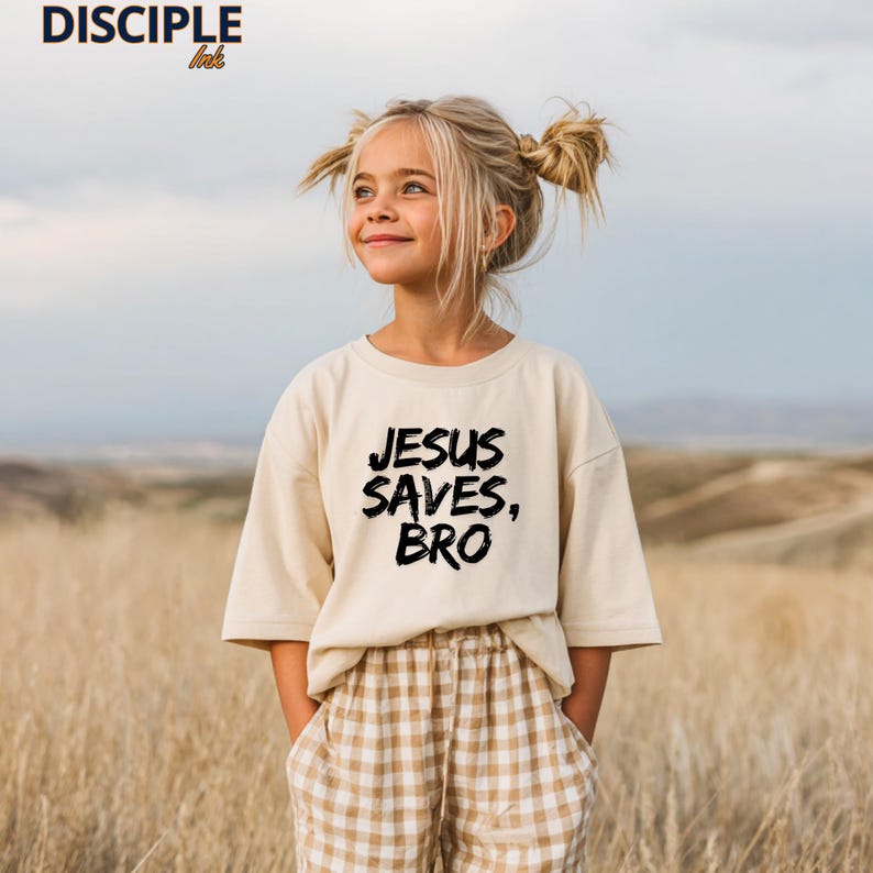 Op de afbeelding: Een beige t-shirt met de tekst "JESUS SAVES, BRO" in zwart, gedragen door een jong persoon. De persoon staat in een veld met hoog gras, met een geruite broek. Het merk "DISCIPLE ink" staat linksboven.