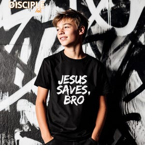 Op de afbeelding: Zwart T-shirt met ronde hals met de tekst "JESUS SAVES, BRO" in witte, versleten letters. Het shirt wordt gedragen door een persoon voor een zwart-witte graffiti-muur. Het merk "DISCIPLE Ink" staat linksboven.