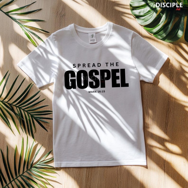 May include: A white short-sleeved t-shirt featuring the black printed text SPREAD THE GOSPEL and MARK 16:15 centered on the chest. The garment has a standard crew neckline and a visible brand tag inside the collar area on the inner back neck.