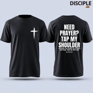 May include: A black short-sleeve t-shirt featuring a white brushstroke cross on the front left chest. The back displays the white text: NEED PRAYER? TAP MY SHOULDER, FOR WHERE TWO OR MORE ARE GATHERED IN MY NAME, THERE AM I WITH THEM. MATTHEW 18:20.