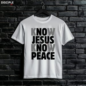 May include: White t-shirt with the text "KNOW JESUS KNOW PEACE" in a stacked layout. The word "JESUS" is in black, while "KNOW" and "PEACE" alternate between gray and black. The shirt is hanging against a dark brick wall.