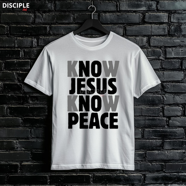 May include: White t-shirt with the text "KNOW JESUS KNOW PEACE" in a stacked layout. The word "JESUS" is in black, while "KNOW" and "PEACE" alternate between gray and black. The shirt is hanging against a dark brick wall.