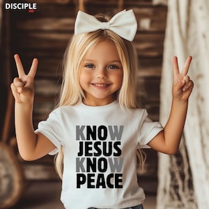 May include: A white t-shirt with the text "KNOW JESUS KNOW PEACE" in black and gray. The child is wearing a white bow and making a peace sign with both hands. The child has blonde hair and is smiling.