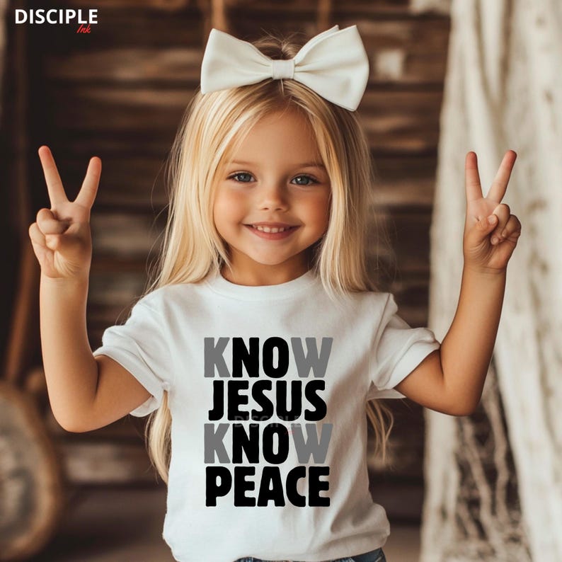 May include: A white t-shirt with the text "KNOW JESUS KNOW PEACE" in black and gray. The child is wearing a white bow and making a peace sign with both hands. The child has blonde hair and is smiling.