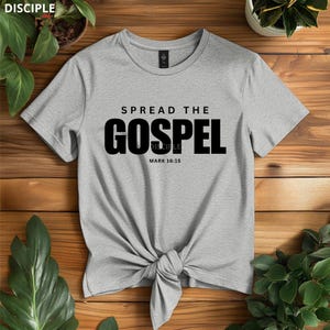 May include: A heather gray short-sleeve t-shirt featuring the black text SPREAD THE GOSPEL and MARK 16:15 printed on the front. The shirt has a crew neckline and is tied in a knot at the bottom center, creating a gathered fabric effect.