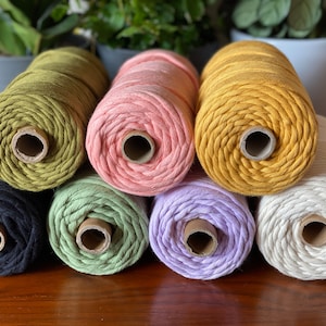 May include: A selection of macrame cord spools in a range of colours, such as olive green, pink, and mustard yellow. The spools are stacked, displaying the texture and colour variations of the cord. Ideal for craft projects, the cord is measured in metres.