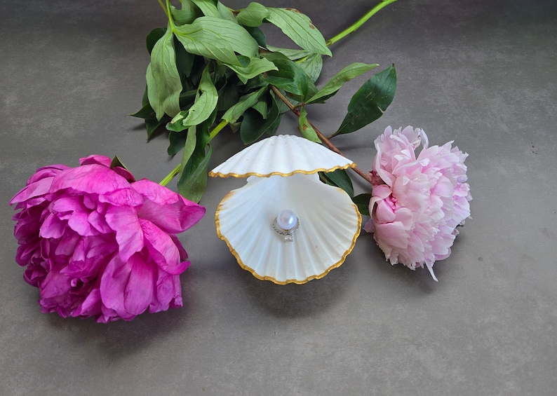 May include: A white seashell with a gold trim, open to reveal a pearl inside. The seashell is surrounded by two pink peonies.