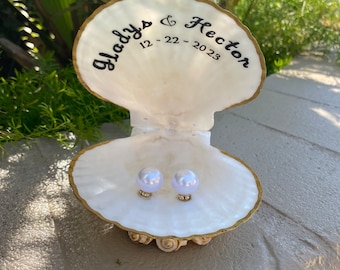 May include: A white seashell with gold trim, open to reveal two white pearl rings. The shell is inscribed with "Gladys & Hector 12-22-2023".