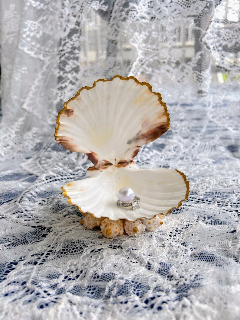 May include: A white seashell with a gold rim, open to reveal a pearl and a ring inside. The seashell is sitting on a white lace tablecloth.