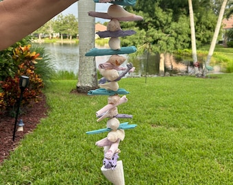 May include: A seashell wind chime featuring driftwood and painted blue and purple accents. The chime is suspended from a white string and crafted from natural materials.