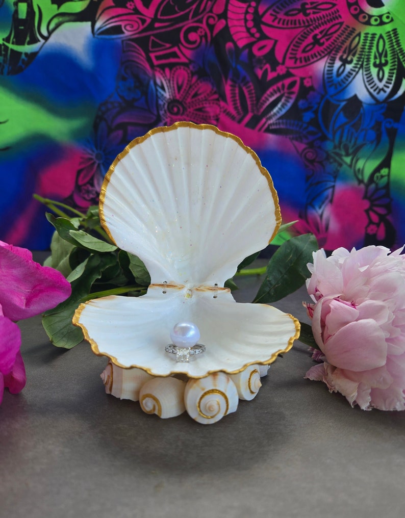 May include: A white seashell ring holder with a gold trim, featuring a pearl and a ring inside. The seashell is sitting on a grey surface with pink flowers in the background.