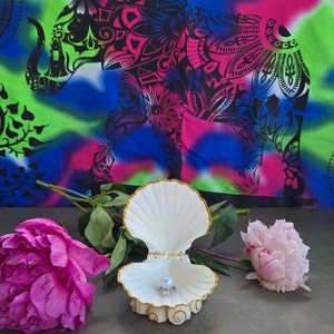 May include: A white seashell ring holder with a gold trim, open to reveal a pearl inside. The holder is sitting on a grey surface with pink flowers on either side.