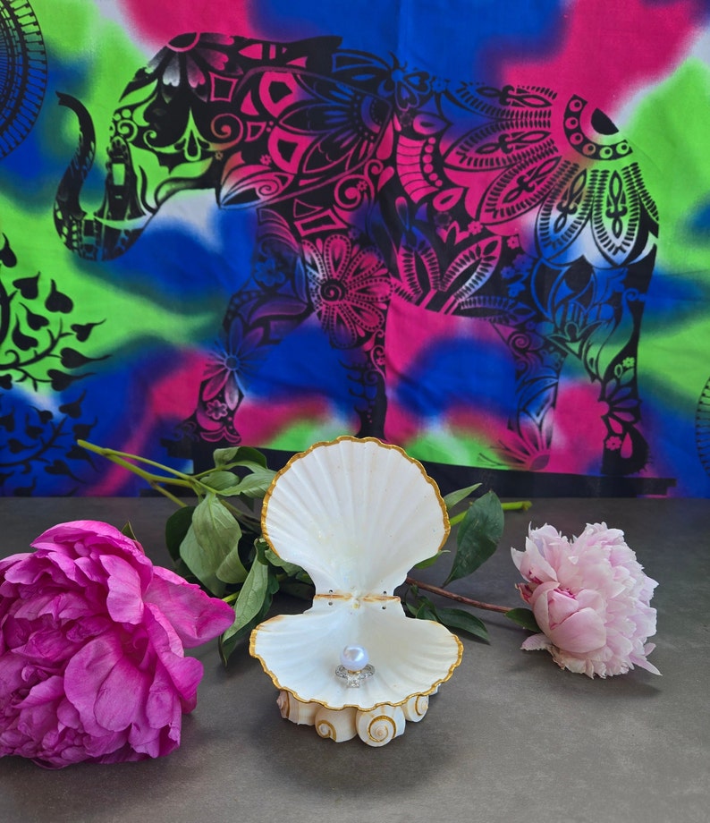 May include: A white seashell ring holder with a gold trim, open to reveal a pearl inside. The holder is sitting on a grey surface with pink flowers on either side.