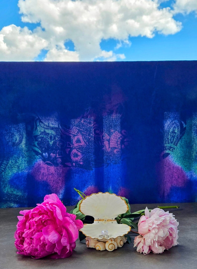 May include: A white seashell ring holder with a pearl ring inside, sitting on a grey surface with two pink peonies on either side. The background is a blue wall with a white cloud in the sky.