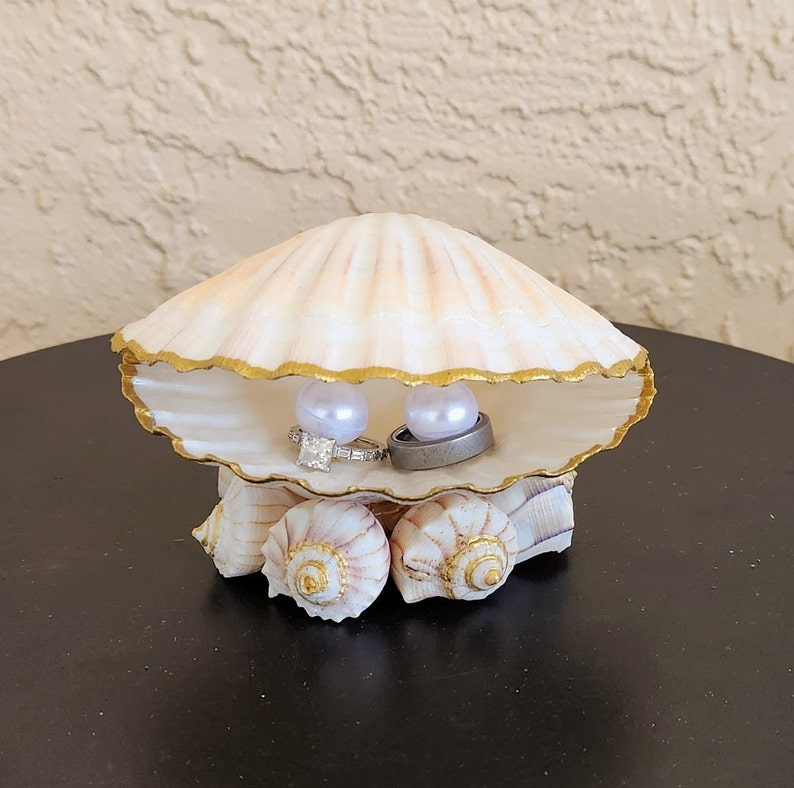 May include: A white seashell ring holder with a gold trim, decorated with two white pearls and two rings. The seashell is sitting on a bed of smaller seashells.