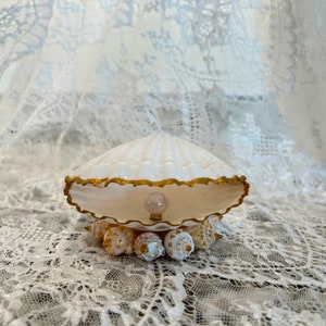 May include: A white seashell with a gold trim, holding a pearl inside. The seashell is sitting on a bed of smaller seashells.