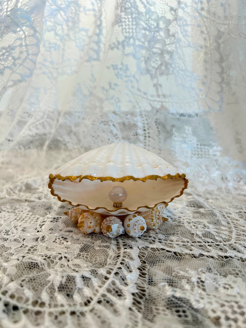 May include: A white seashell with a gold trim, holding a pearl inside. The seashell is sitting on a bed of smaller seashells.