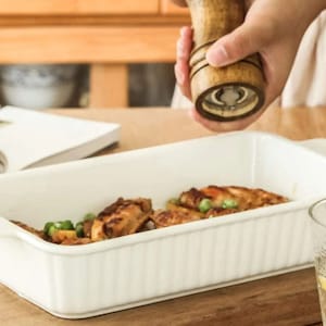 May include: A white rectangular baking dish with a fluted rim, filled with roasted chicken wings and green peas. A wooden pepper mill is being used to season the dish.