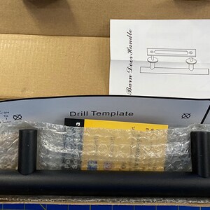 May include: Black barn door handle with a drill template and instructions. The handle is made of metal and has a simple design. The template is printed on a white sheet of paper and includes measurements in inches.