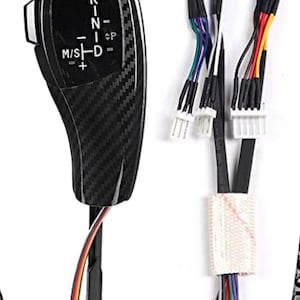 May include: A black carbon fiber gear shift knob with a silver gear pattern and the letters "R-N-D-M/S-P" on the side. The knob is connected to a black wire with a multi-colored wire coming out of the bottom. The image also shows a set of black wires with blue connectors and white connectors.