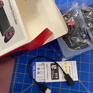 May include: A black and red video game controller with a USB cable and a warranty card. The card has a gift card for $10 on Amazon.com.